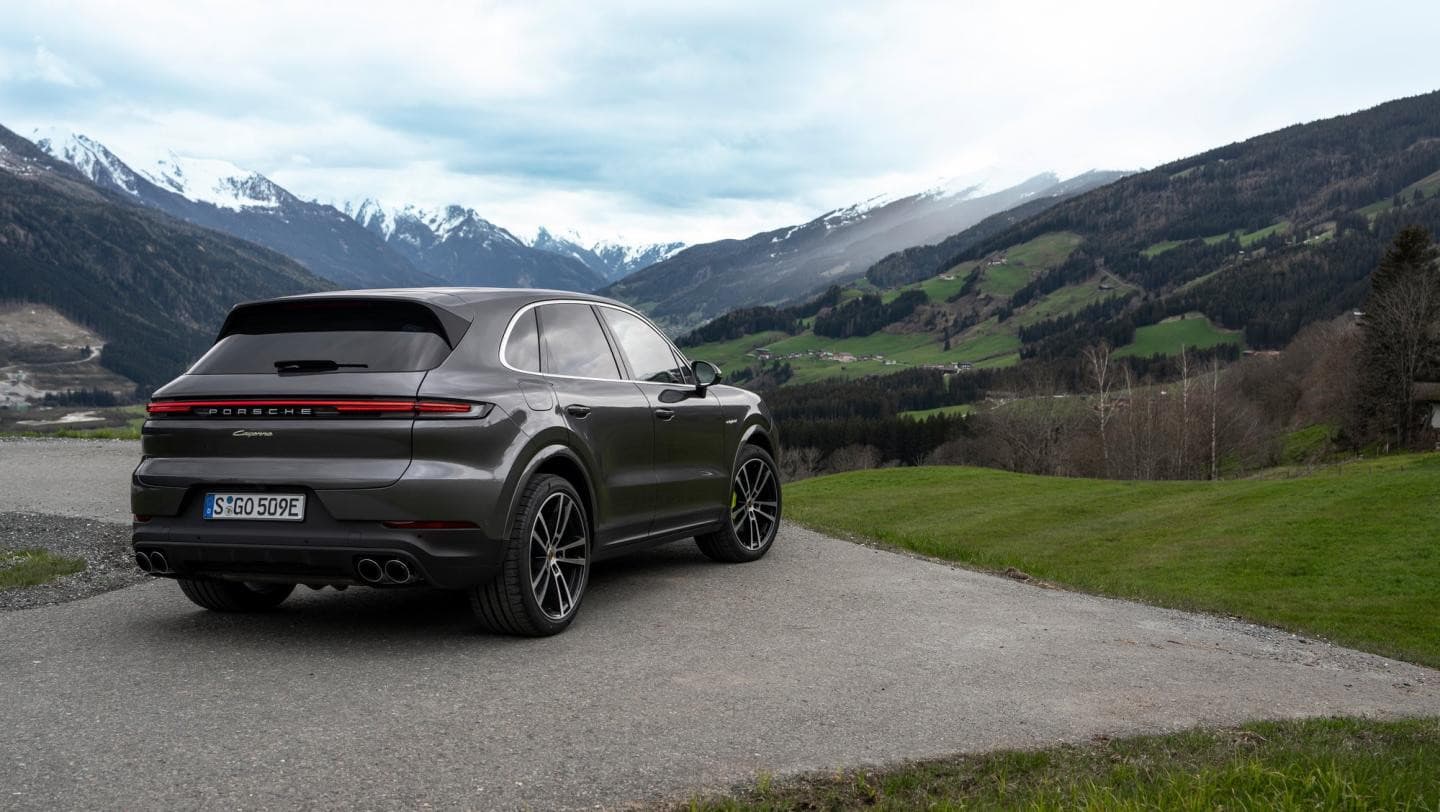 Grey Porsche Cayenne E-Hybrid, rear three-quarter view, with a mountain valley behind it